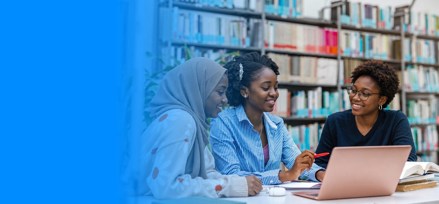 Three learners smiling while working together on a laptop in a library