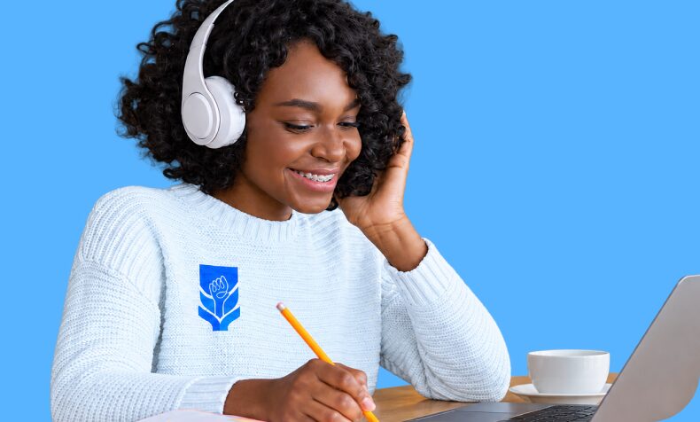 A learner wearing headphones takes notes beside her laptop